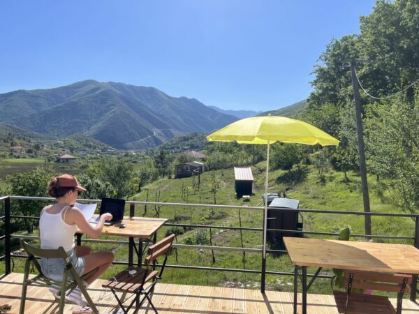 student zit achter een laptop op een houten terras met gekleurde parasol, een groene tuin en bergen op de achtergrond met blauwe lucht - scriptiereizen Albanië